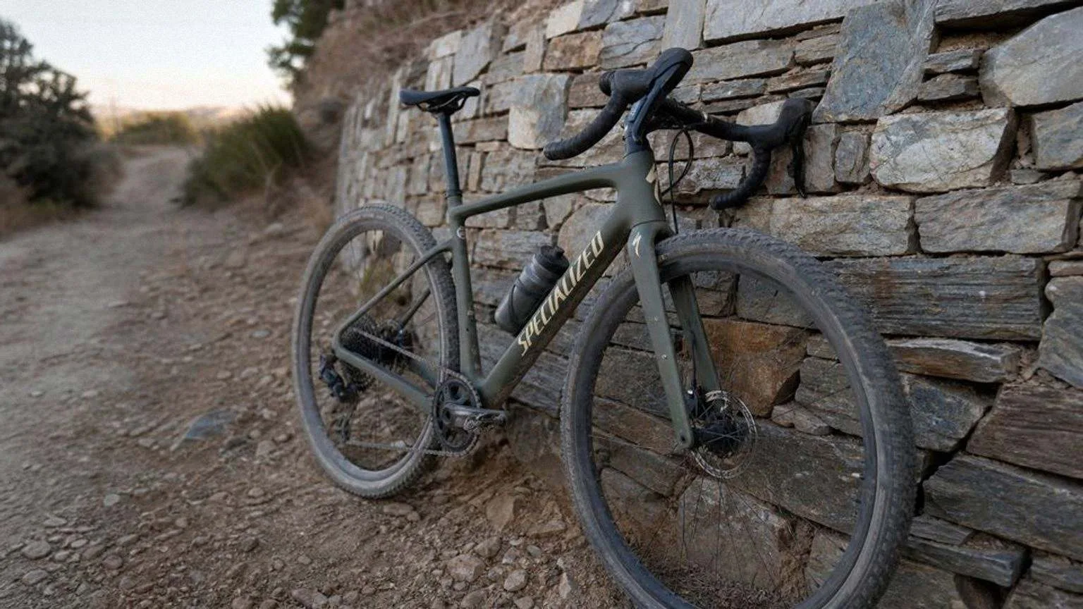 Bicicleta de gravel con cuadro verde oliva apoyada en un muro de piedra seca junto a un camino de tierra, con cubiertas anchas llenas de polvo y luz baja de media tarde.