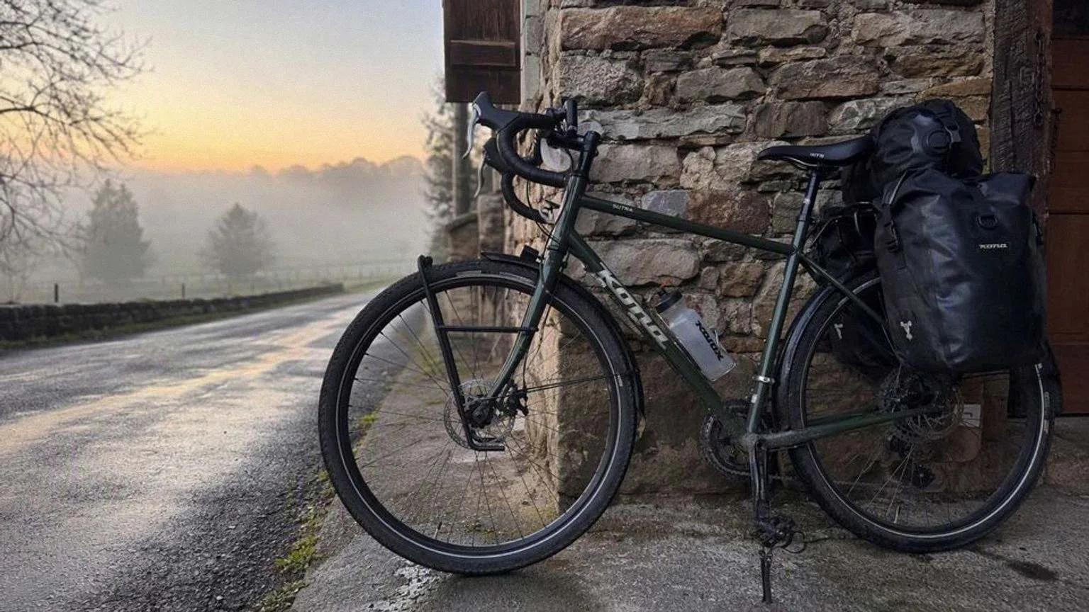 Bicicleta de cicloturismo de acero con portabultos delantero y trasero, dos alforjas cargadas y guardabarros, apoyada en un muro de piedra al amanecer junto a una carretera secundaria mojada.