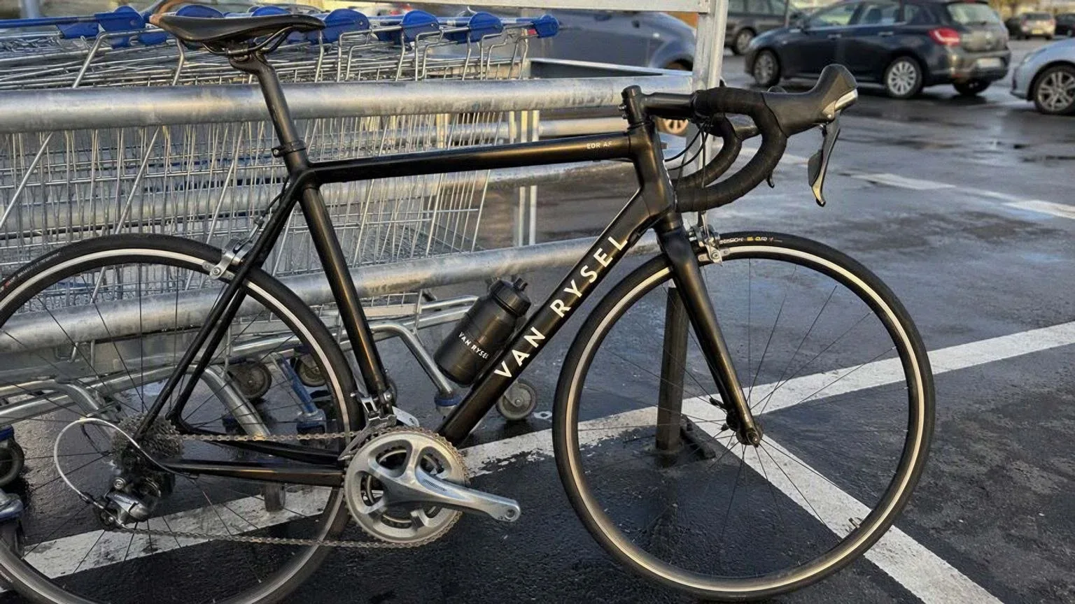 Bicicleta de carretera de aluminio negro con manillar de curva y frenos de llanta, apoyada en un aparcamiento de superficie con el asfalto mojado al final de la tarde.