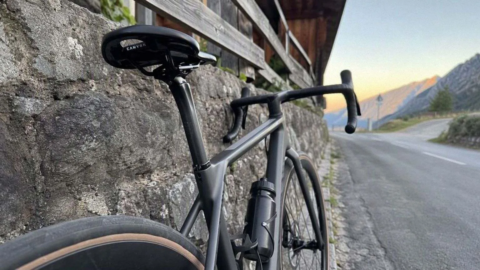 Bicicleta de carretera de carbono gris antracita apoyada contra un muro de piedra en una carretera secundaria de montaña, con la tija de carbono y el sillín en primer plano al amanecer.