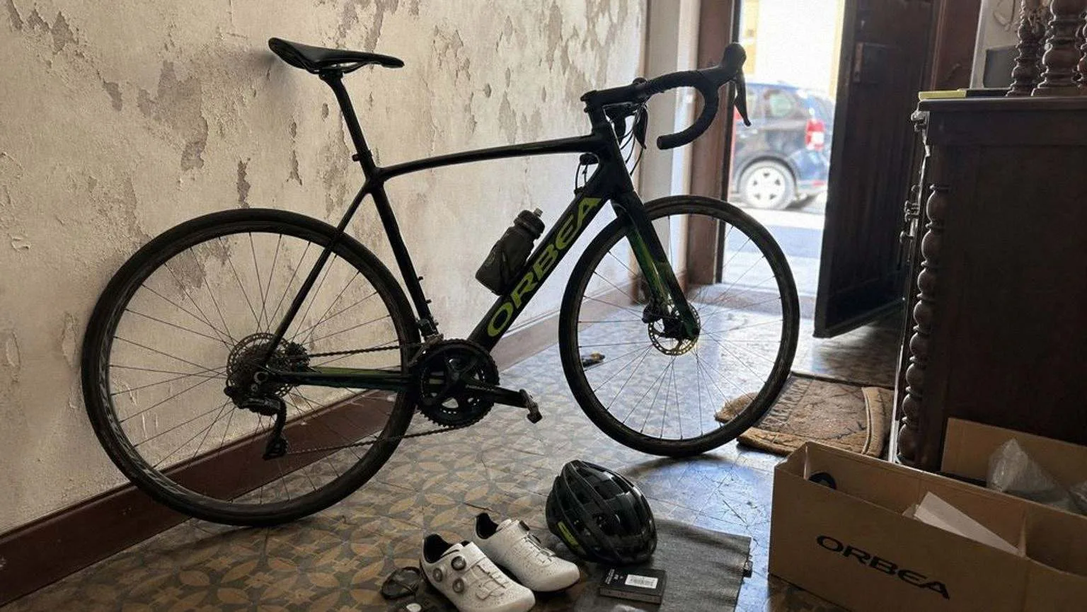 Bicicleta de carretera de carbono negro con detalles verdes, frenos de disco y cubiertas de 28 milímetros, aparcada en el portal de un piso.
