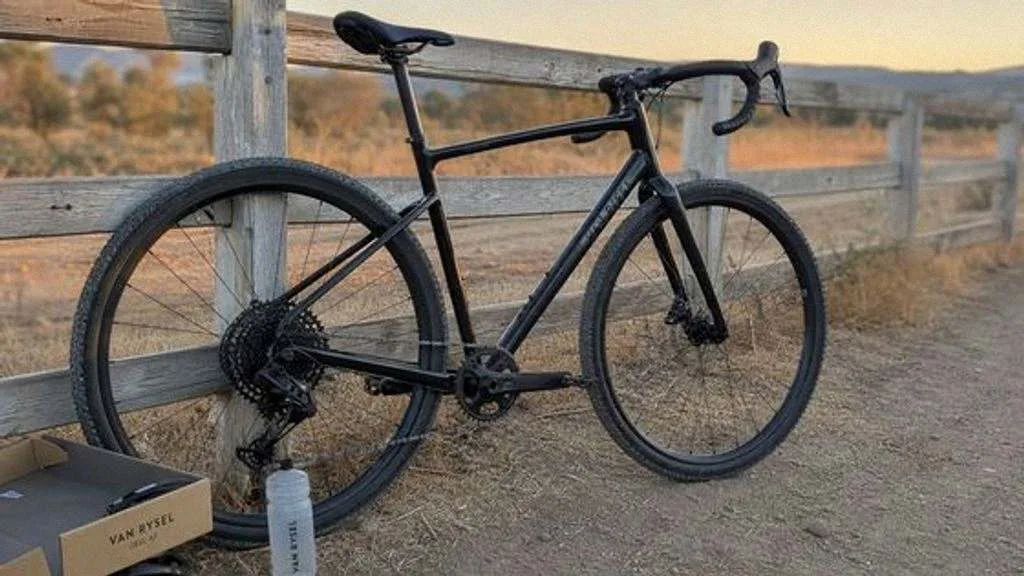 Una gravel de aluminio negra apoyada en una valla de madera junto a un camino de tierra, con cubiertas anchas y manillar de curva.