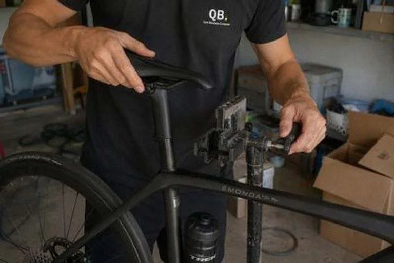 Una mano sujeta una bicicleta de carretera por el sillín mientras la otra abre la mordaza de un caballete de taller en un garaje con cajas y una manguera en el suelo.