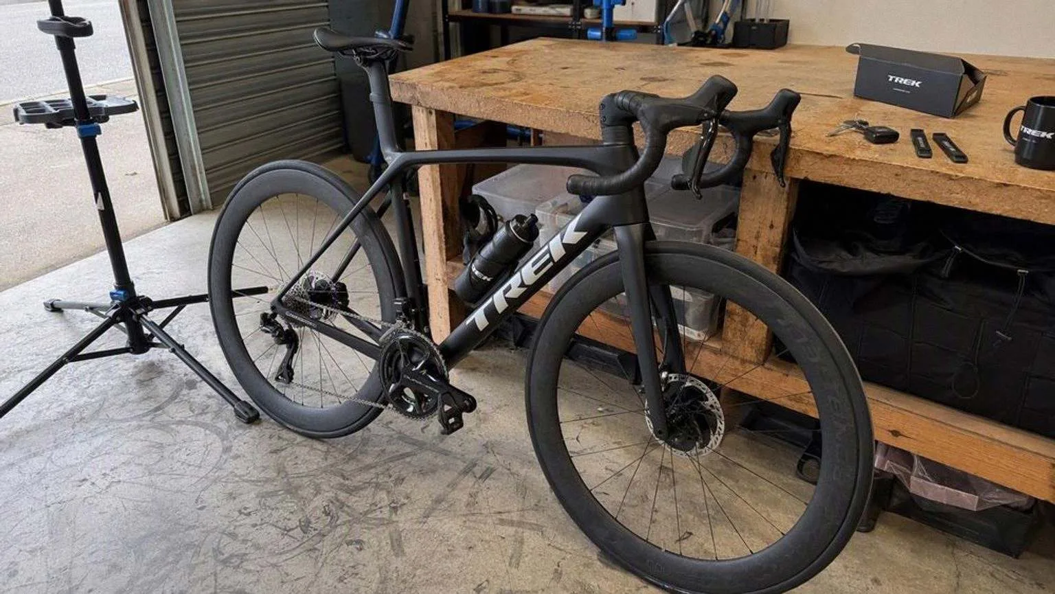Bicicleta de carretera de carbono negra con ruedas de perfil alto, apoyada en el banco de un taller junto a un caballete de reparación y unos desmontables.