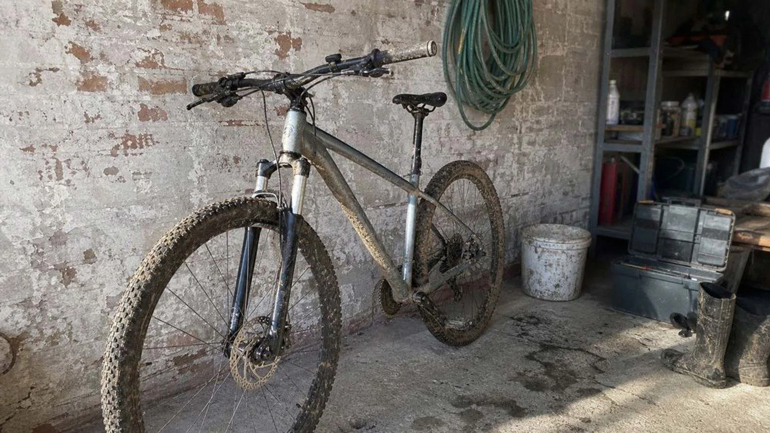 Bicicleta de montaña rígida con barro seco en el cuadro y en las cubiertas, apoyada en una pared de ladrillo junto a una manguera, un cubo y una caja de herramientas.